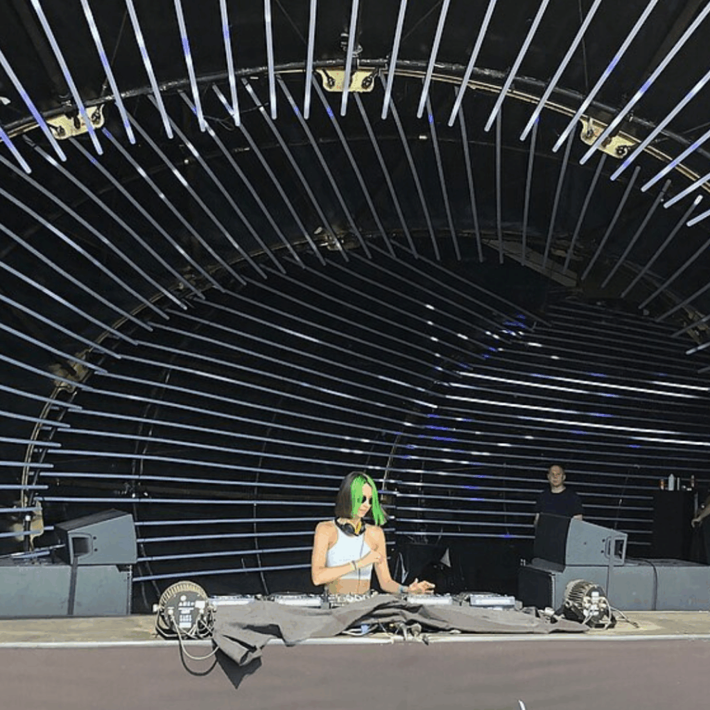 DJ Miss Monique en live derrière ses platines sous une grande structure métallique en arc, scène de musique électronique.