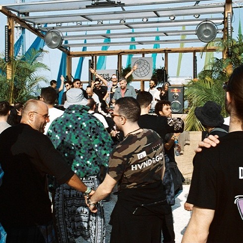 Foule devant un DJ set en plein air sur la plage, sous une structure décorée avec des rubans colorés