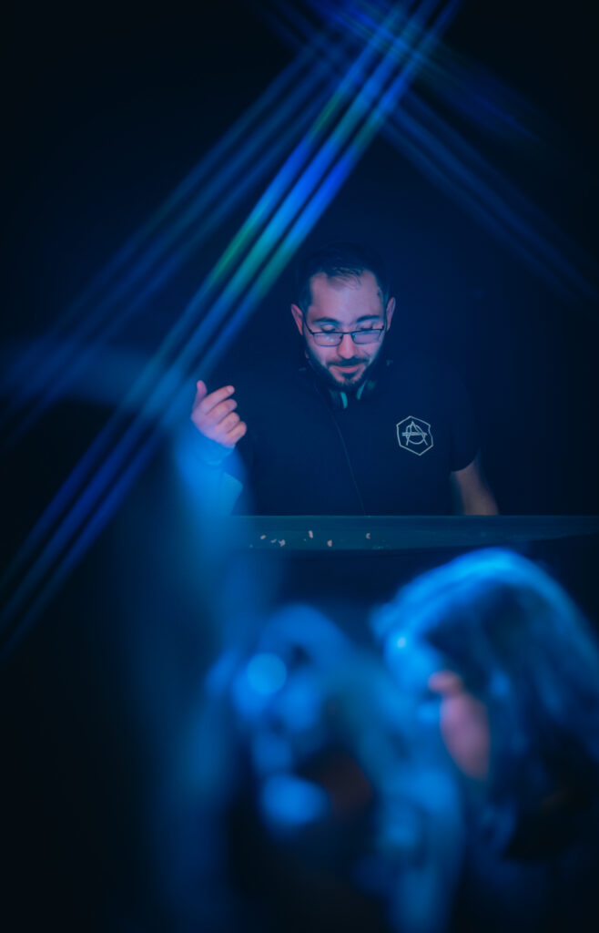 DJ AY3L mixe derrière son booth avec des faisceaux de lumière bleue et le public flou au premier plan