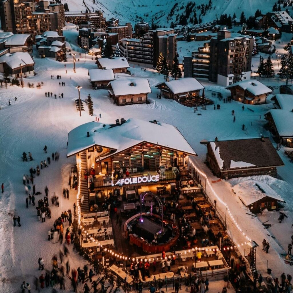 Vue aérienne nocturne de la station des Deux Alpes enneigée avec un grand bâtiment éclairé au premier plan.